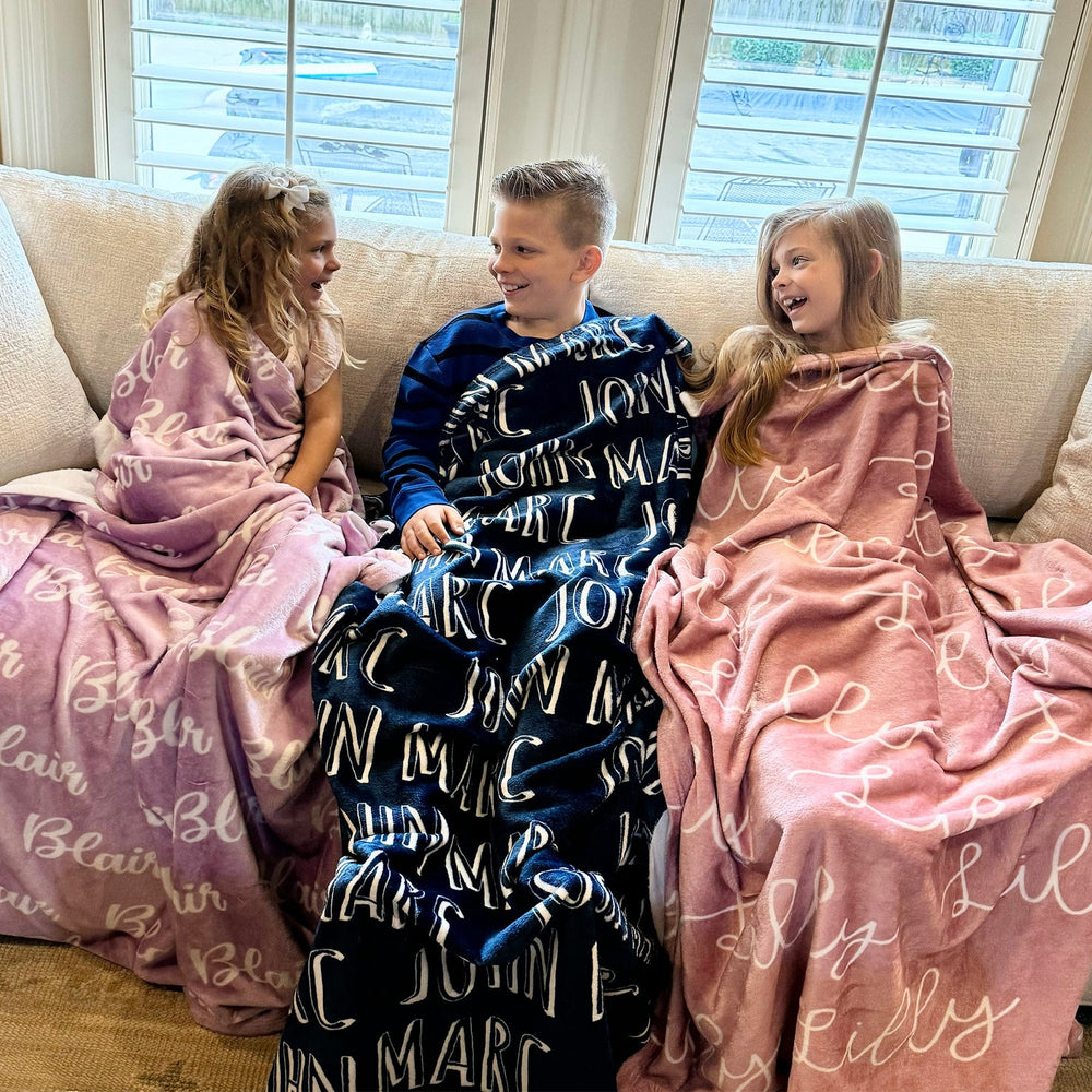 3 kids enjoying their personalized name blankets.  The blankets are pink, navy and lilac in color with 3 different fonts.